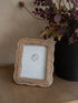 A small rectangular wicker picture frame with a scalloped edge, standing on a woven surface beside a black vase filled with dried dark foliage.