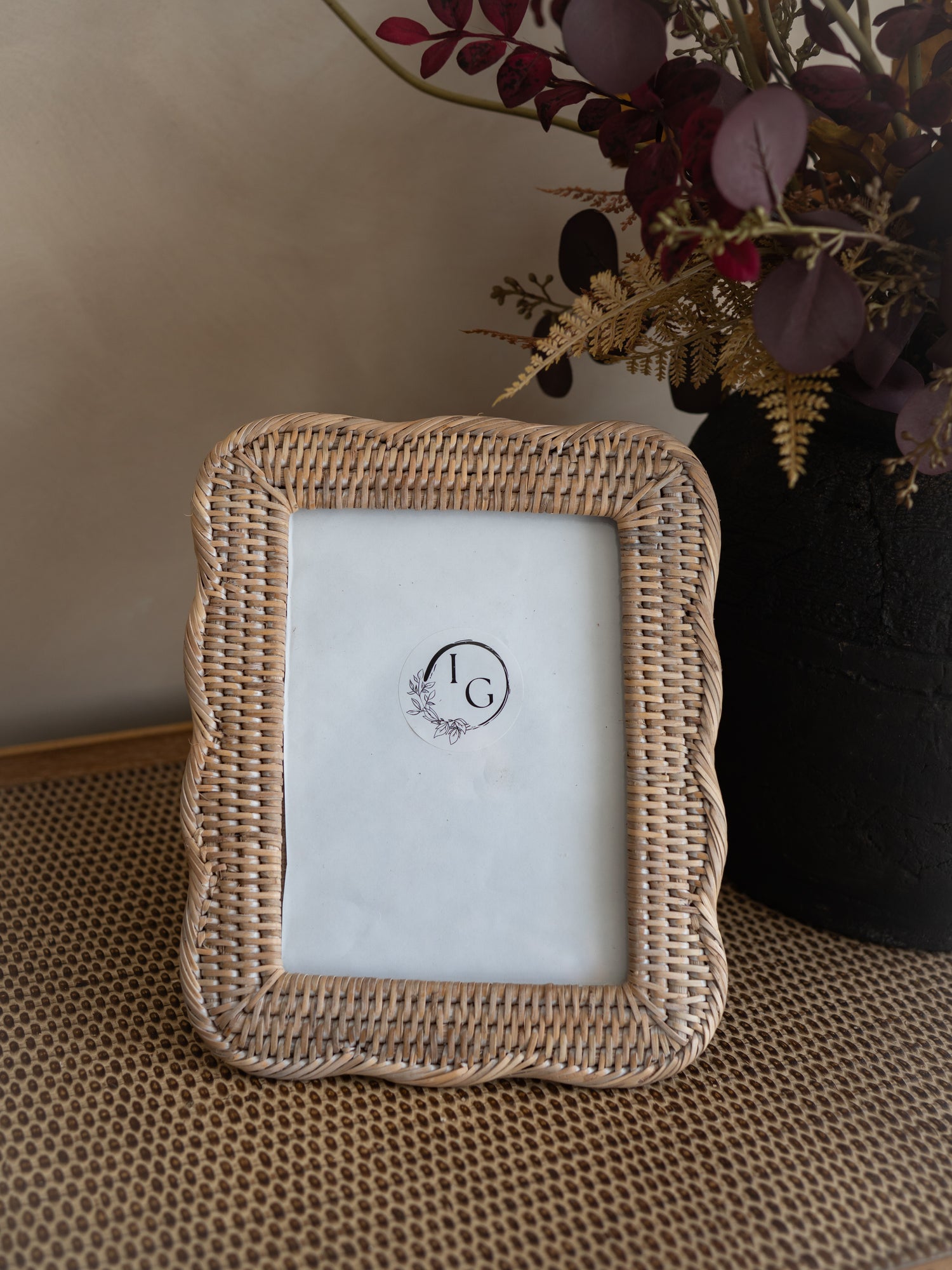 A rectangular woven rattan picture frame with a scalloped edge, standing on a textured surface next to a black vase with dried foliage.