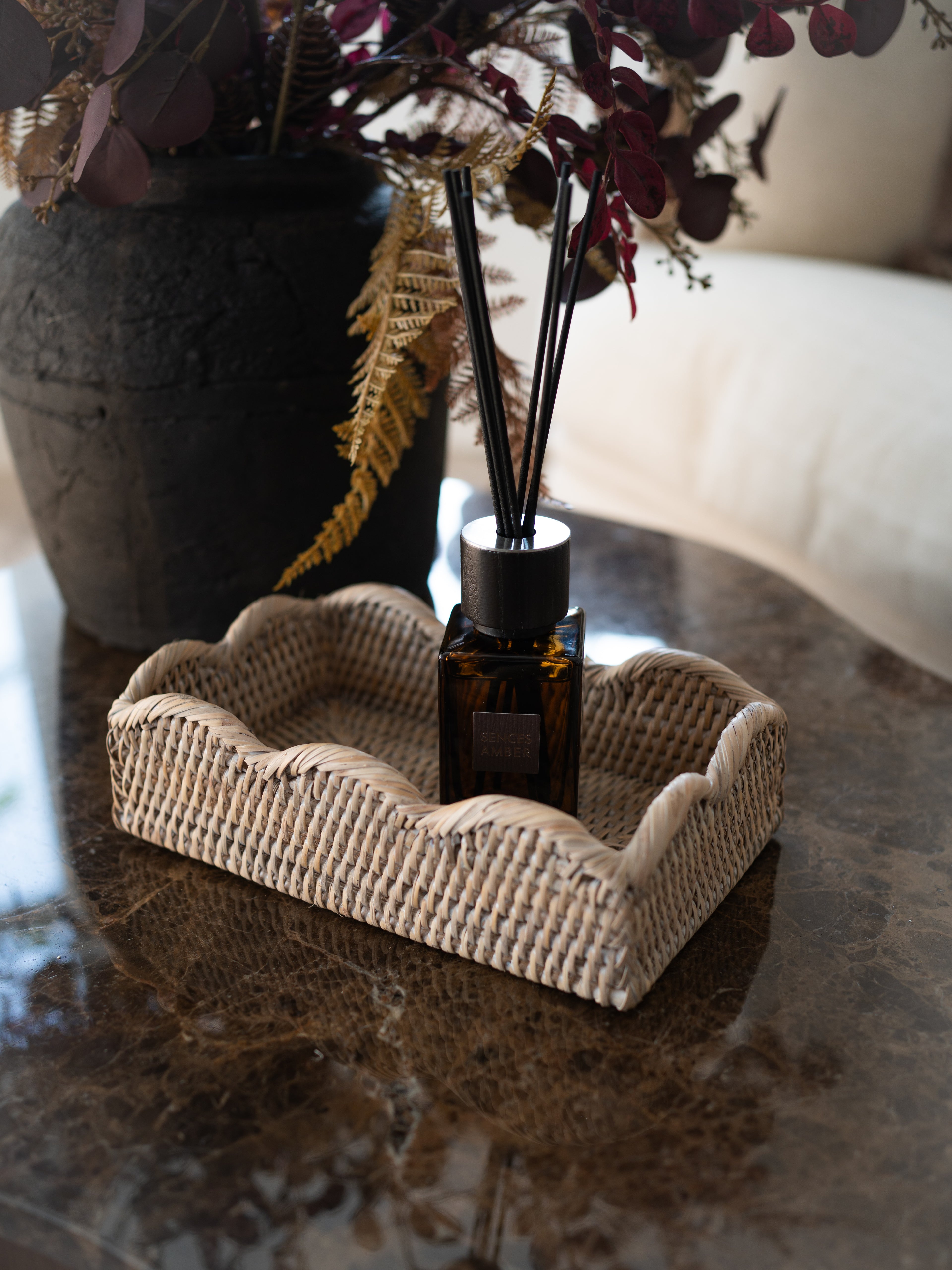 A reed diffuser bottle with black reeds sits in a woven white washed rattan tray with scallop edge detail on a polished dark marble table, with a large dark planter and dried foliage in the background.