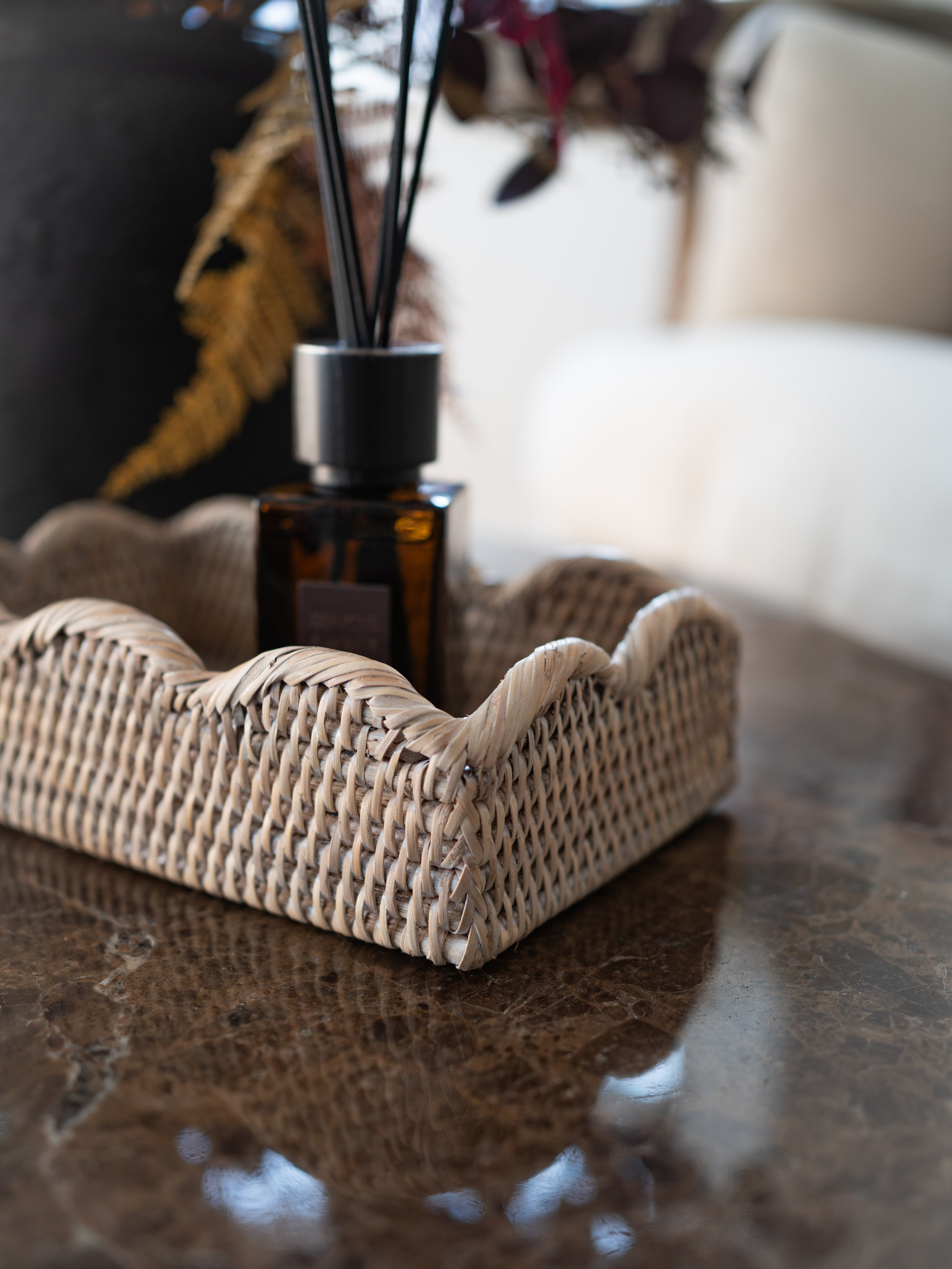 A woven rattan tray with scallop edge holding a dark amber glass reed diffuser bottle with black reeds, placed on a polished brown marble surface in a softly blurred indoor setting.