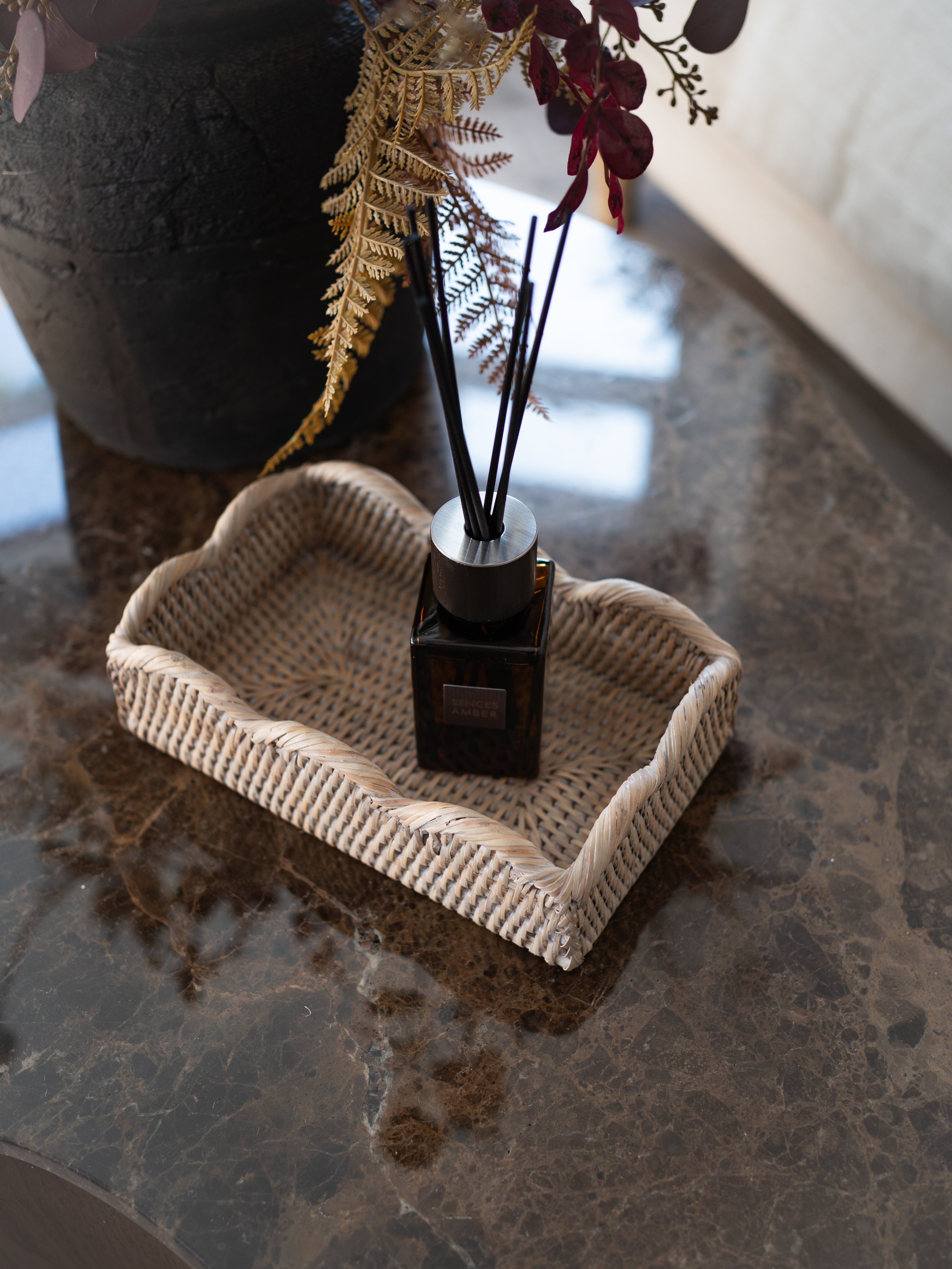 A reed diffuser bottle with black reeds sits in a woven white washed scallop rattan tray on a polished dark marble surface, with dried flowers and a dark vase in the background.