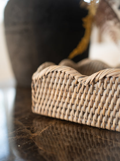 A close-up of a woven rattan fiber basket or tray with a wavy top edge, resting on a dark reflective surface with a blurred indoor background.