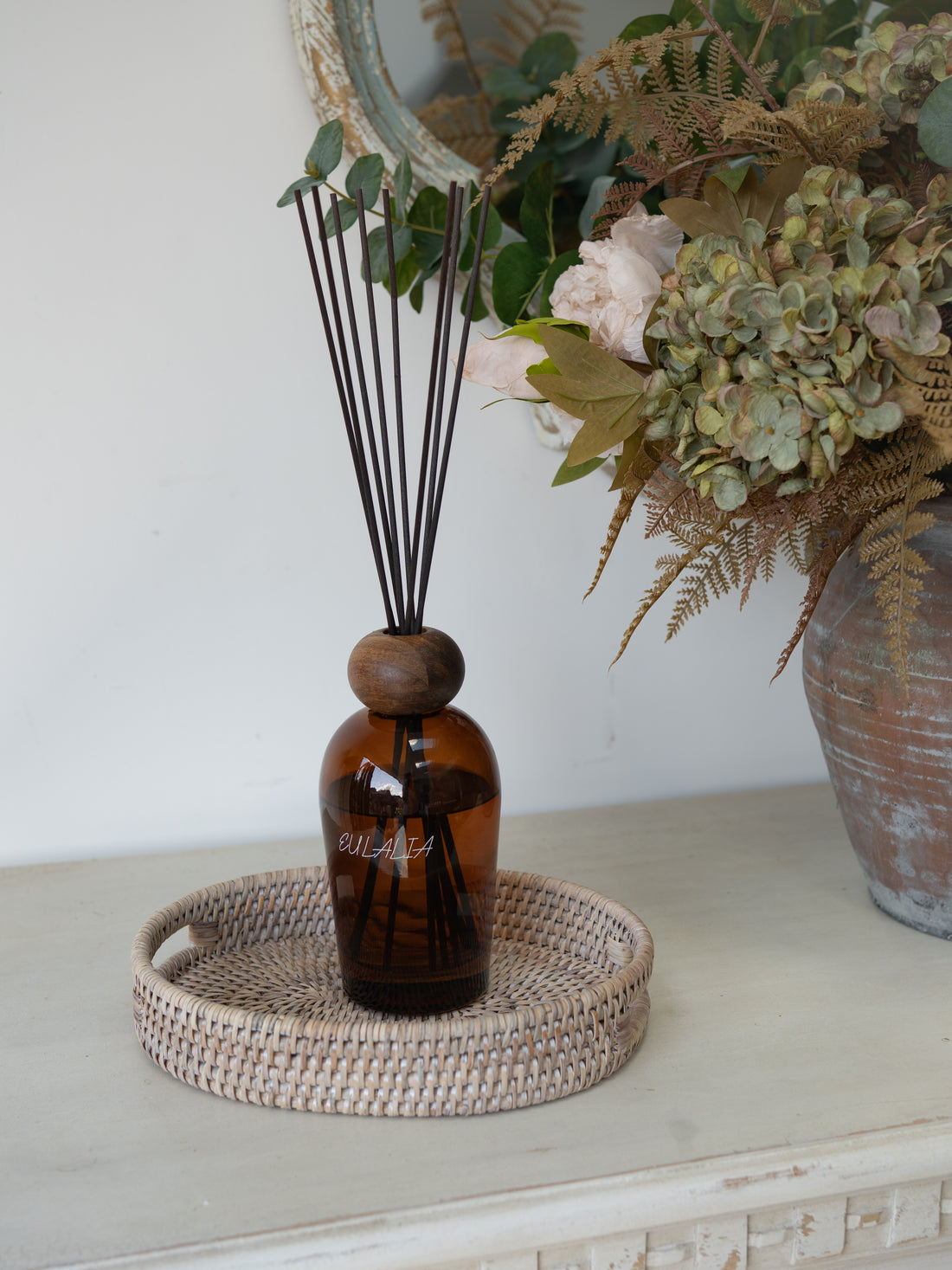 White Washed Rattan Small Oval Tray with brown bottle reed diffuser in and faux autumn flowers in rustic vase in the background