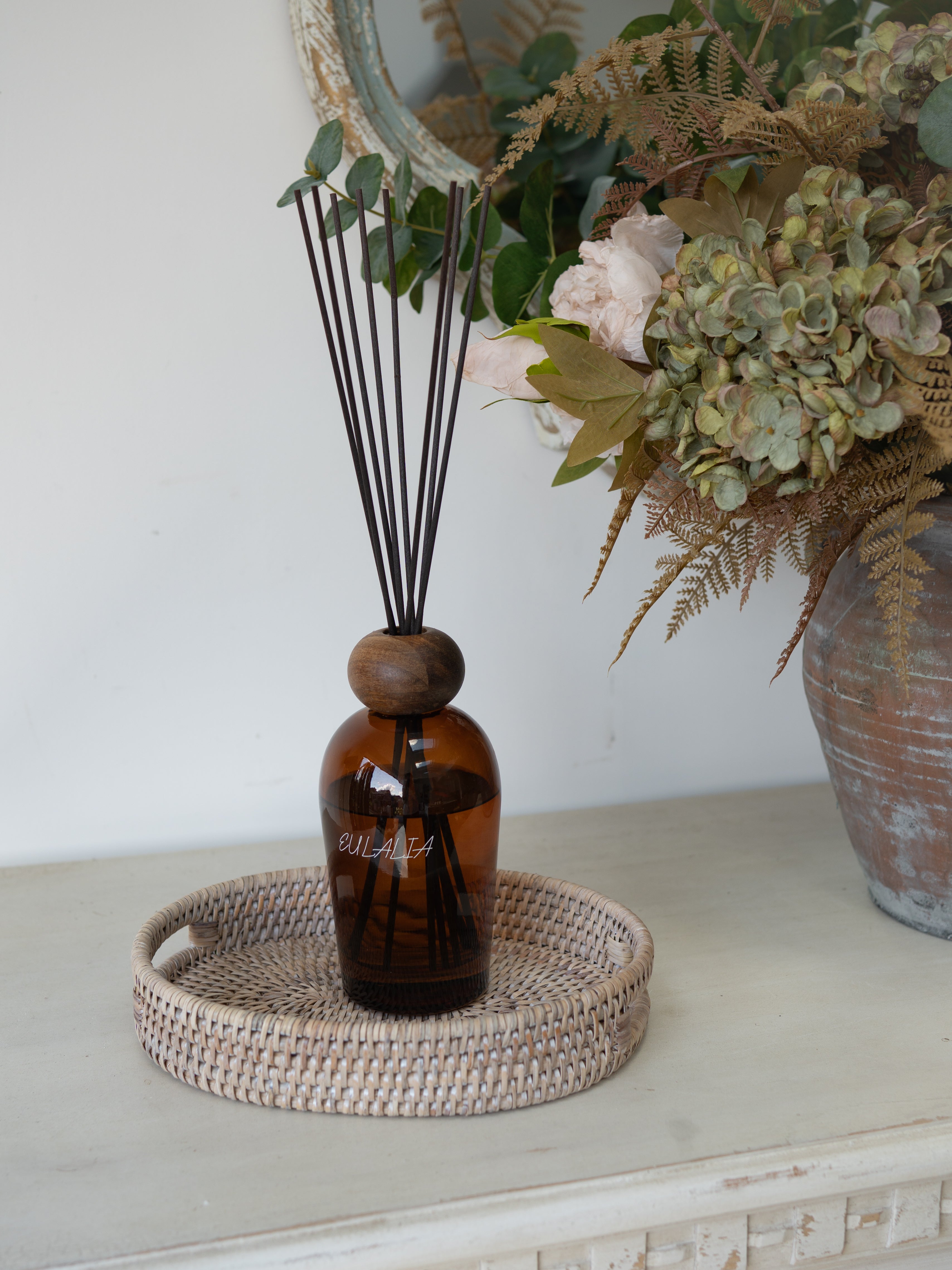 White Washed Rattan Small Oval Tray with brown bottle reed diffuser in and faux autumn flowers in rustic vase in the background