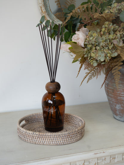 White Washed Rattan Small Oval Tray with brown bottle reed diffuser in and faux autumn flowers in rustic vase in the background