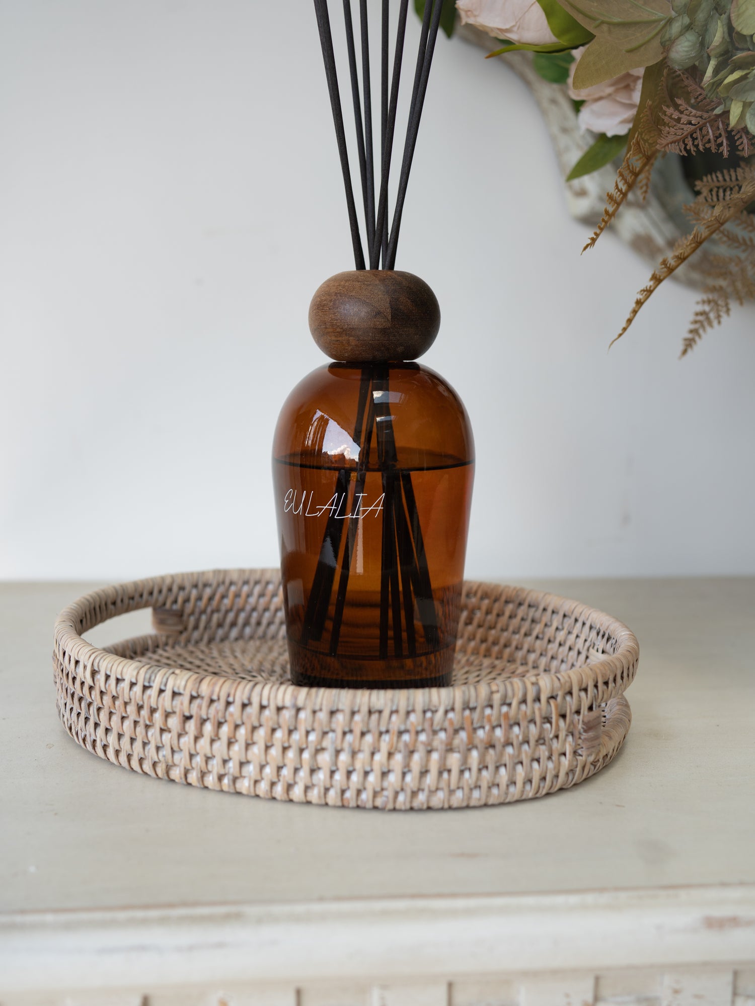 Amber glass reed diffuser bottle with a round wooden stopper and black reeds, displayed on a woven  rattan oval tray against a light neutral background.