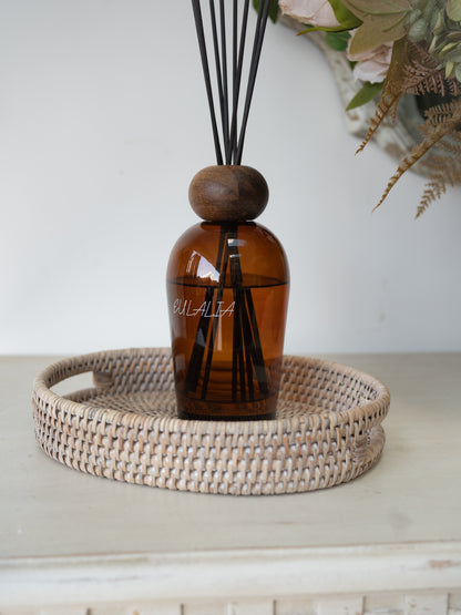 Amber glass reed diffuser bottle with a round wooden stopper and black reeds, displayed on a woven  rattan oval tray against a light neutral background.
