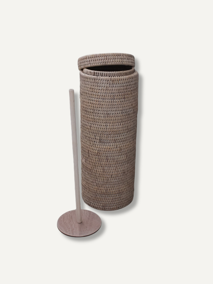 A tall cylindrical woven basket with a matching lid, shown next to a slim light wood floor stand on a plain off-white background.