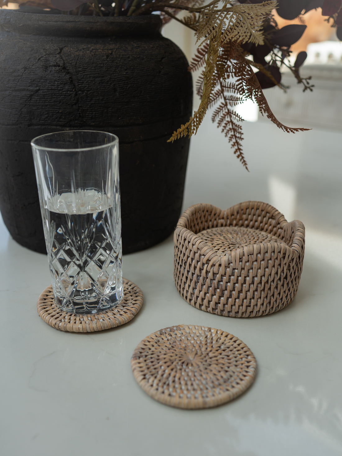 Natural woven seagrass coasters in different shapes, including a round coaster in the foreground and two scalloped-edge coasters in the background, shown on a light surface with a glass of water and a black vase.