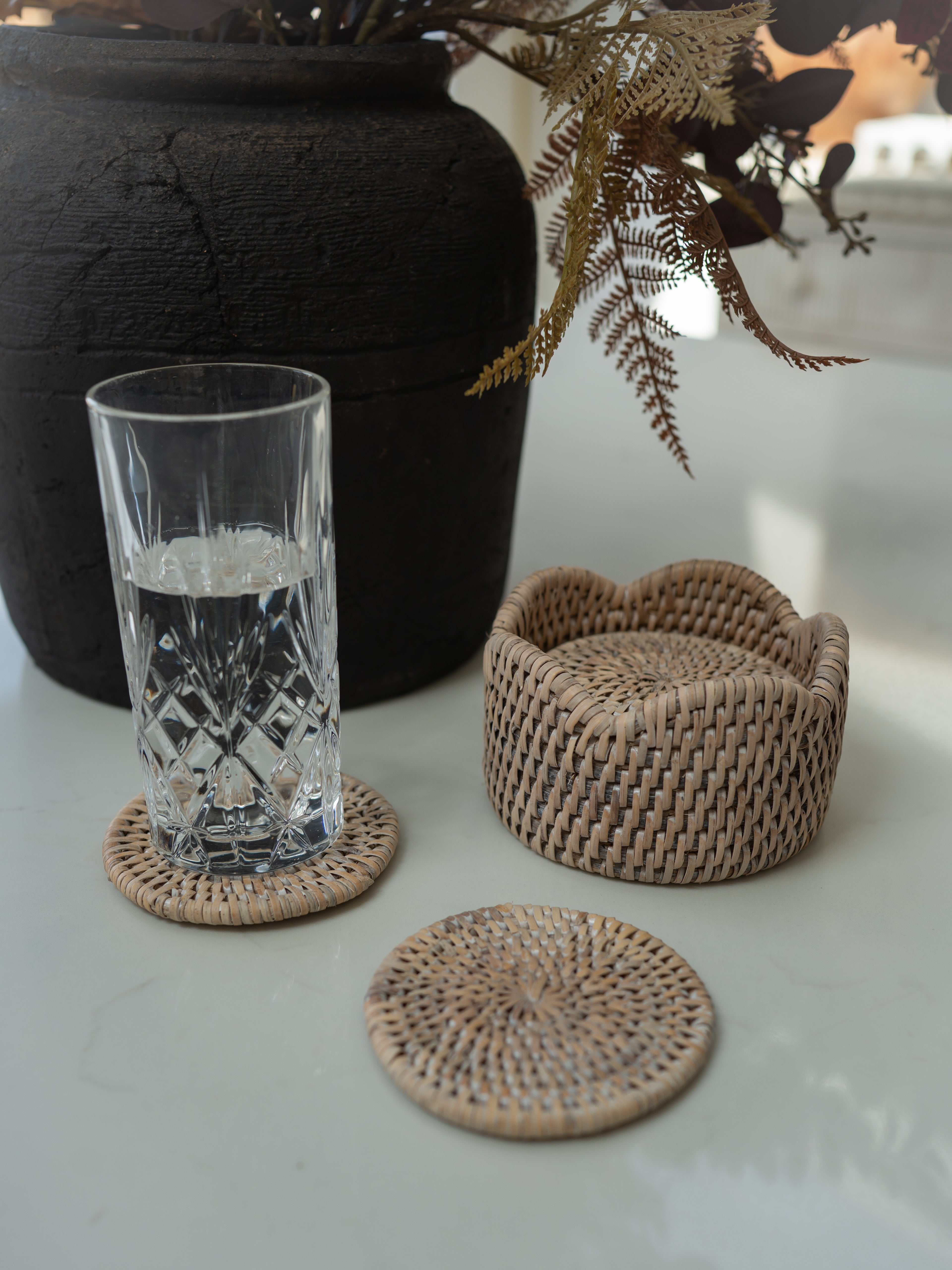 Natural woven seagrass coasters in different shapes, including a round coaster in the foreground and two scalloped-edge coasters in the background, shown on a light surface with a glass of water and a black vase.