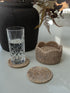Natural woven seagrass coasters in different shapes, including a round coaster in the foreground and two scalloped-edge coasters in the background, shown on a light surface with a glass of water and a black vase.