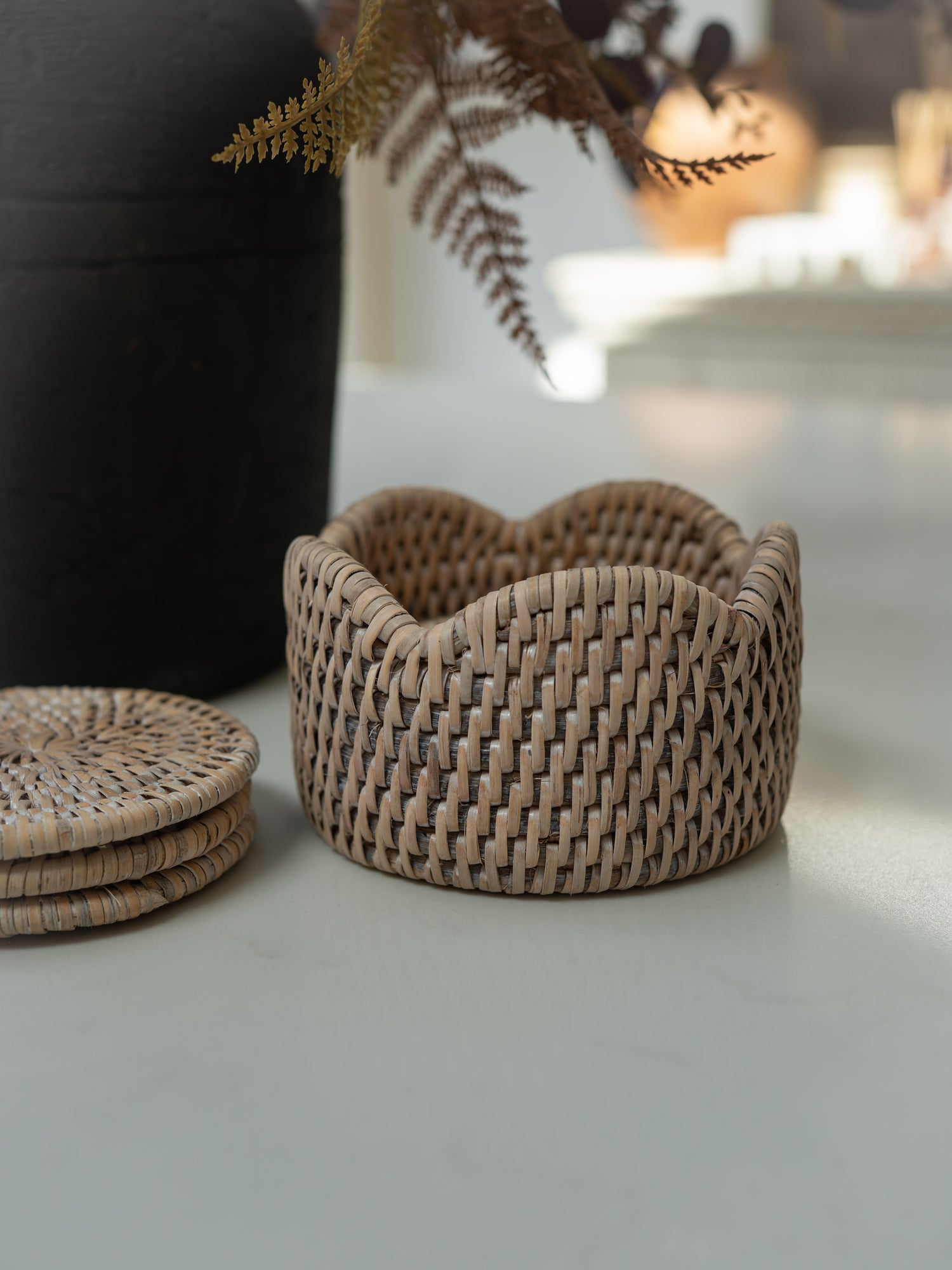 A small woven coaster holder with a scalloped rim in a natural tan color, styled on a light gray surface next to a stack of matching coasters and a dark vase with dried ferns.