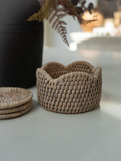A small woven coaster holder with a scalloped rim in a natural tan color, styled on a light gray surface next to a stack of matching coasters and a dark vase with dried ferns.