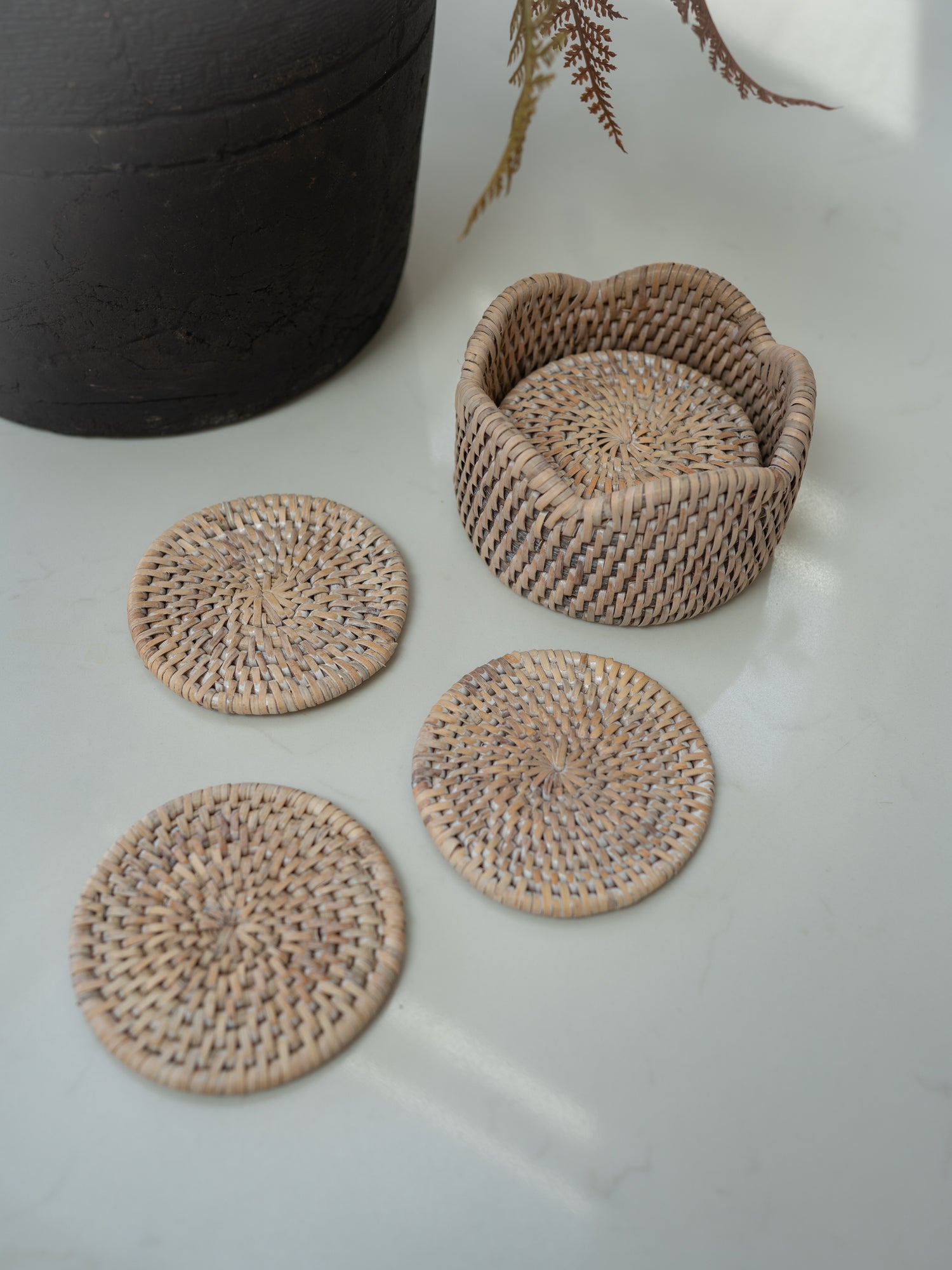 A set of natural woven rattan coasters and holder, including a small flower-shaped basket and four round coasters, arranged on a light gray surface next to a dark ceramic pot.