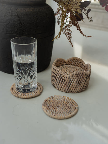 Natural woven rattan coasters and a matching round box on a light tabletop, with a clear drinking glass and dried ferns in the background.