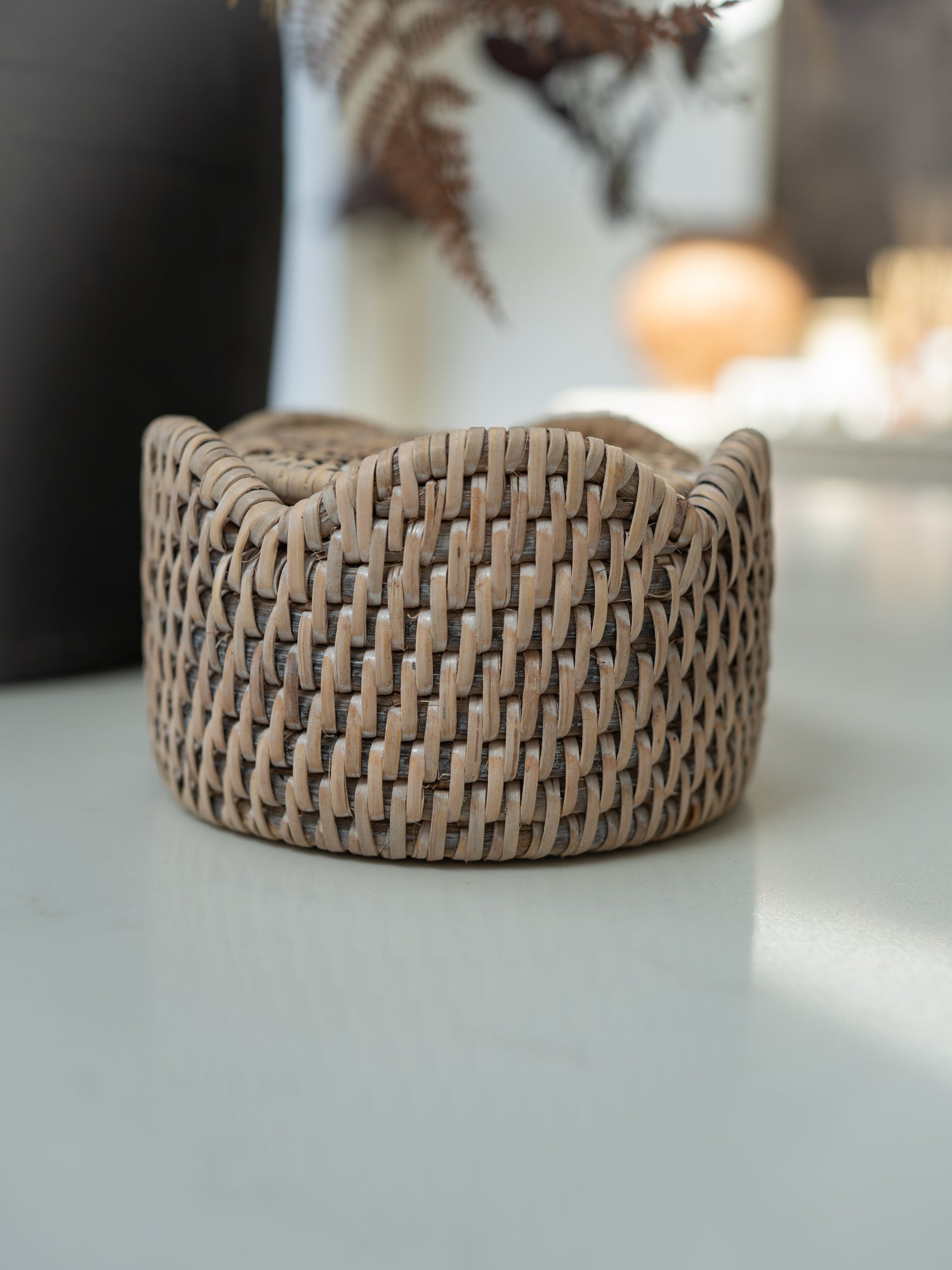 A small woven coaster holder with a rounded, tiered shape in a natural light brown rattan or wicker material, sitting on a light gray surface with a blurred background.