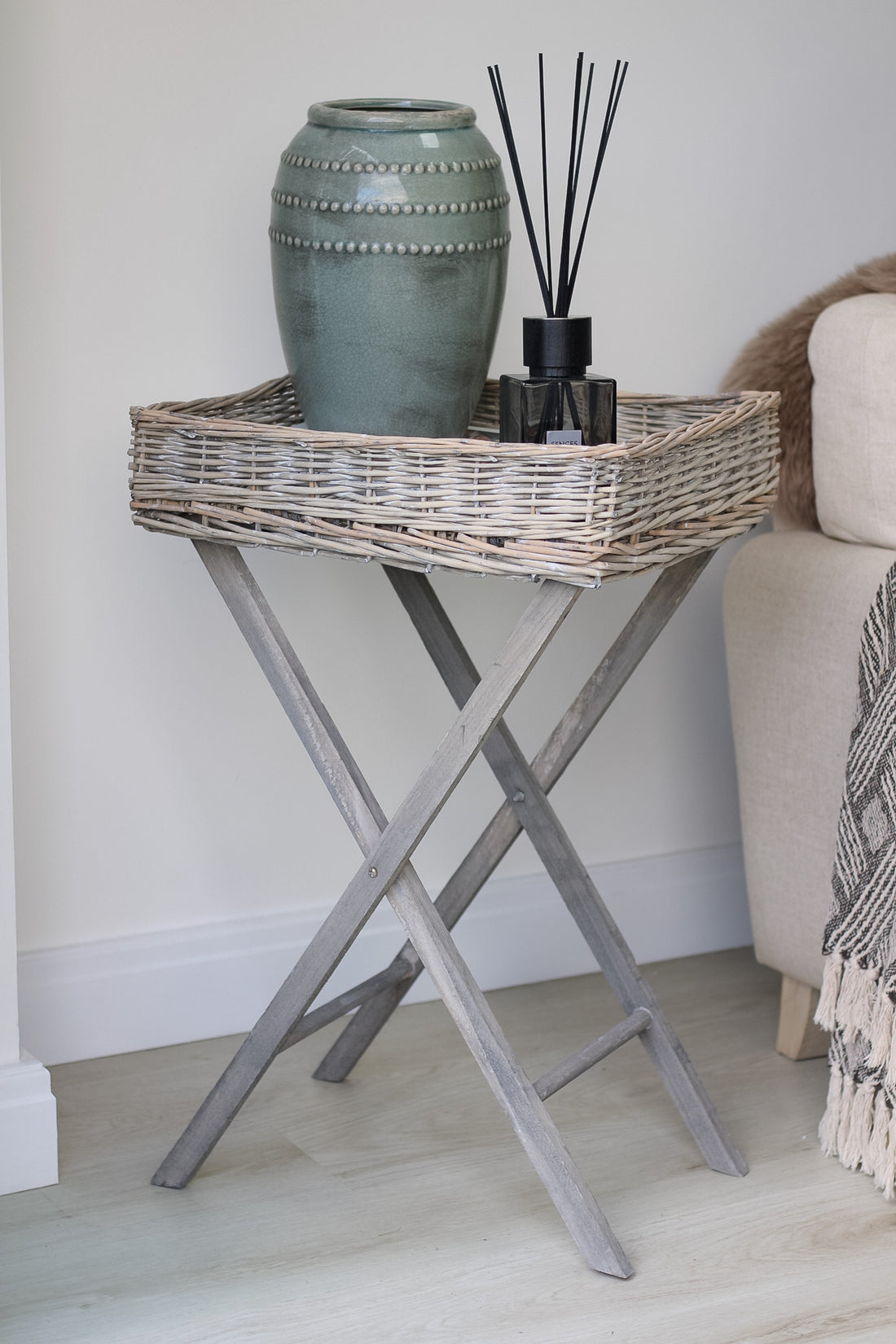 A wicker butler tray table with a gray frame and legs, and a beige wicker tray. It is displayed in a room setting with decorative items on top.