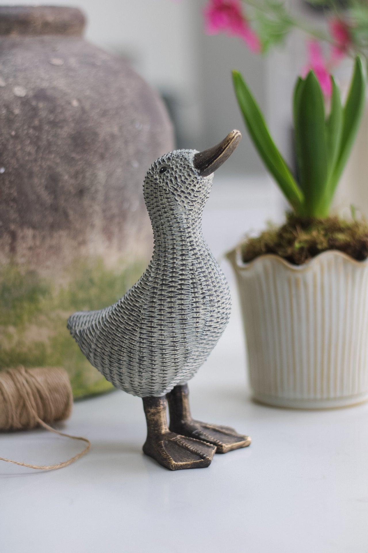 A decorative wicker-style resin duck with a bronze metal base, positioned on a white surface with a potted plant in the background.