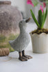 A decorative wicker-style resin duck with a bronze metal base, positioned on a white surface with a potted plant in the background.