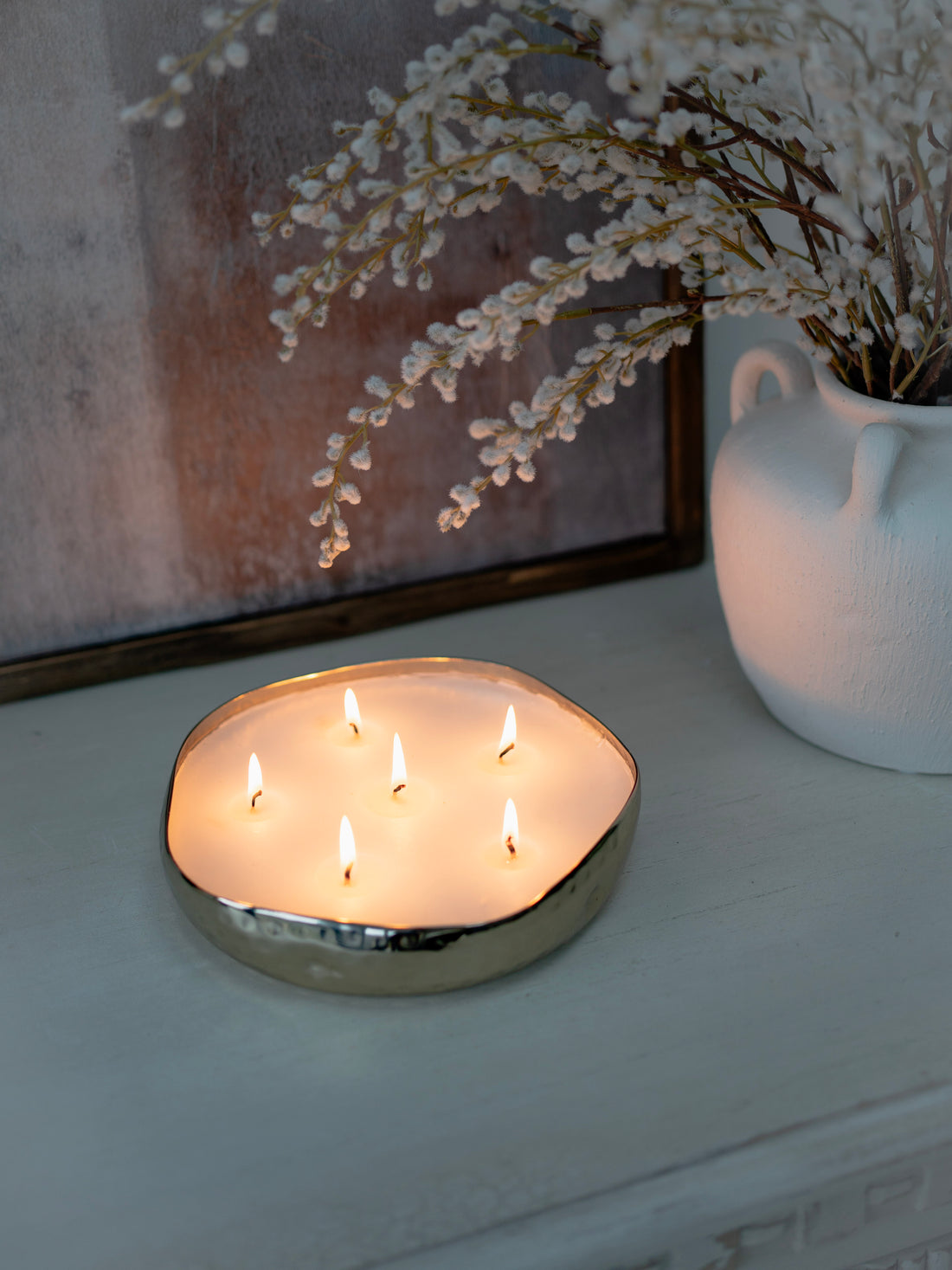 Round metal bowl candle holder with multiple candles on a surface next to a vase with white flowers.