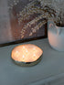 Round metal bowl candle holder with multiple candles on a surface next to a vase with white flowers.