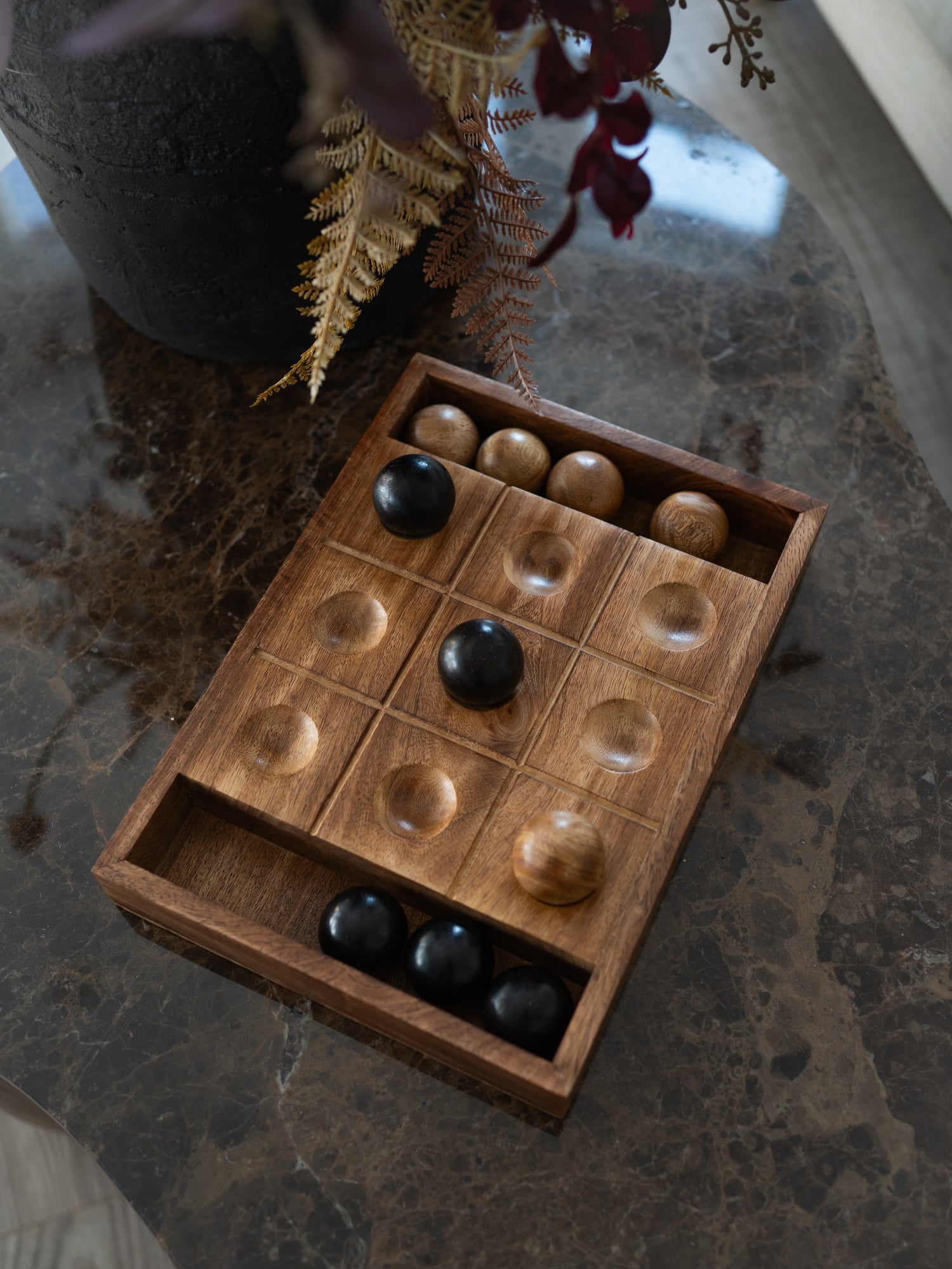 A wooden tic tac toe game board with round wooden and black checkers, displayed on a dark marble tabletop next to a dark vase with dried foliage.