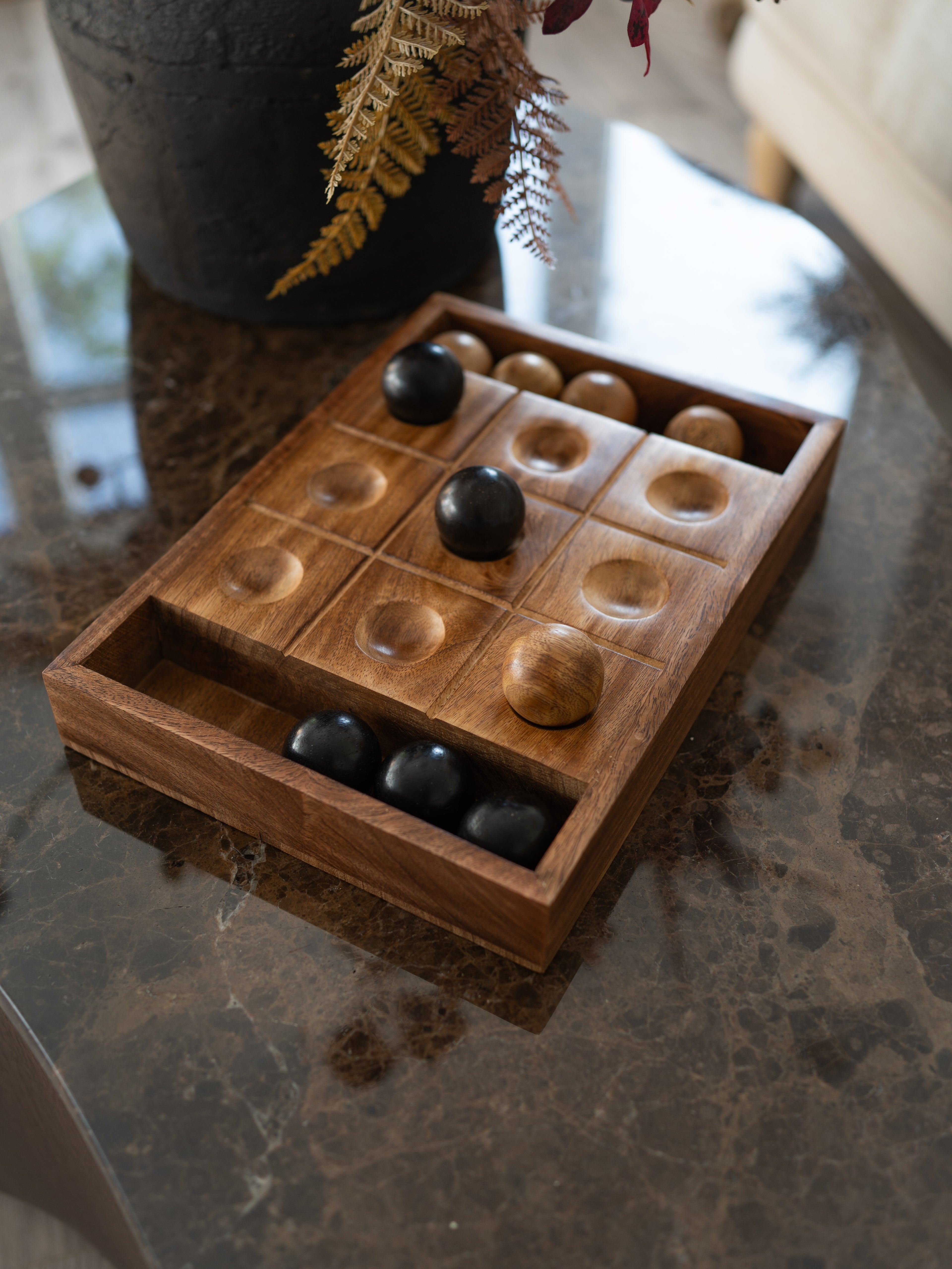 A wooden tic tac toe board game with carved compartments and polished black and natural wood balls, displayed on a dark marble tabletop.