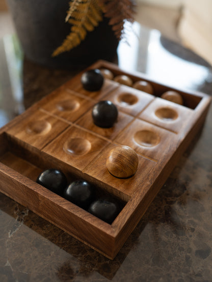 A wooden tic tac toe game board with six compartments and several polished wooden and black stones, photographed on a dark stone countertop in a real indoor setting.