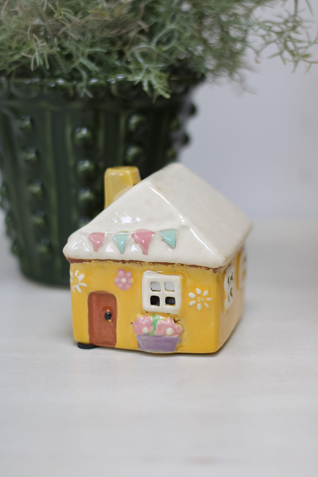 Small ceramic house-shaped candle holder with a white roof and yellow body, decorated with colourful flags and flowers, on a light background.