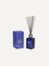 A reed diffuser bottle with a faceted cobalt blue glass body, silver cap, and black reeds, shown next to its matching blue packaging box on a light beige background.