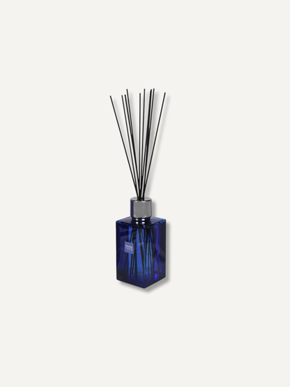 A reed diffuser with a faceted cobalt blue glass bottle and black reeds, set against a solid off-white background.