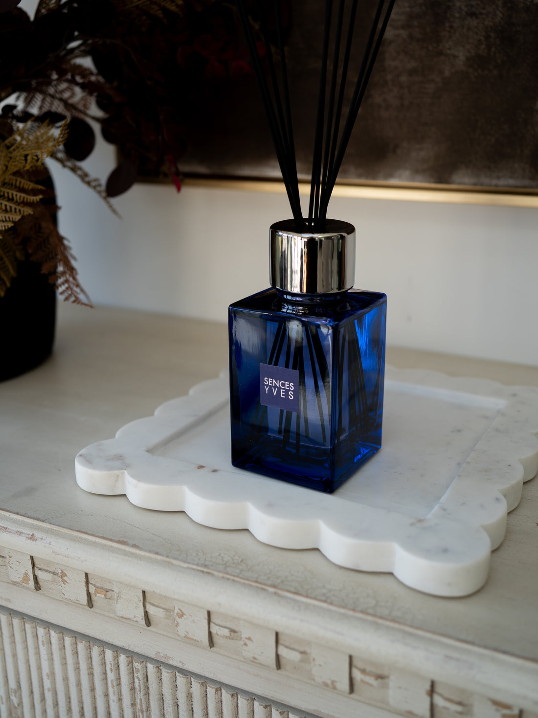 A cobalt blue glass reed diffuser bottle with a silver cap and black reeds, resting on a white scalloped tray on a distressed white console table.