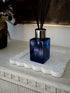 A cobalt blue glass reed diffuser bottle with a silver cap and black reeds, resting on a white scalloped tray on a distressed white console table.