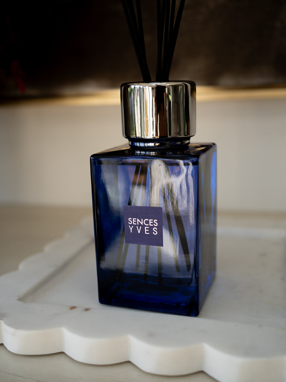A Sences Yves reed diffuser with a square cobalt blue glass bottle, a shiny silver cap, and black reeds, resting on a white scalloped tray.