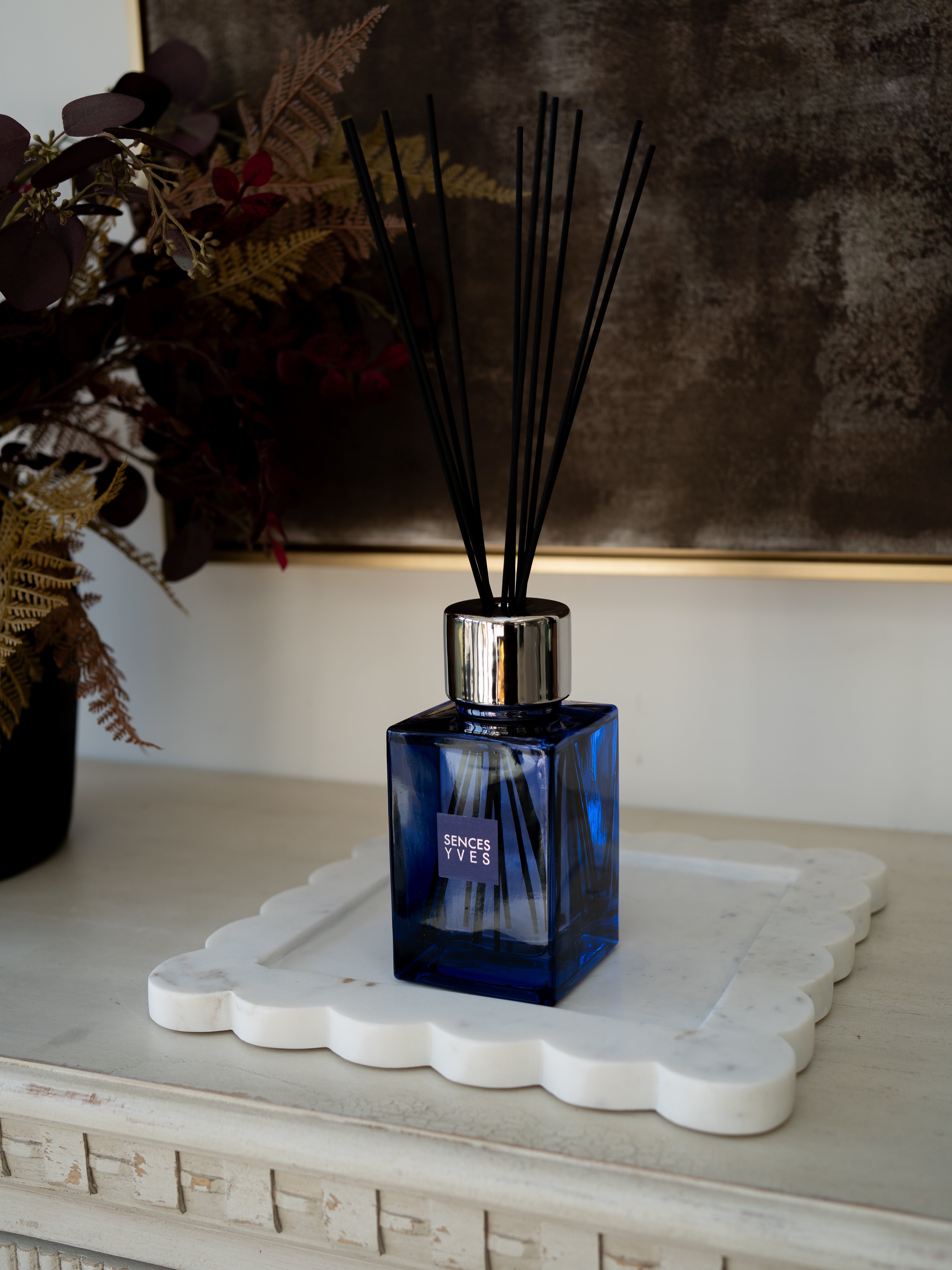 A blue glass reed diffuser bottle with a silver cap and black reeds, placed on a white scalloped tray on a light console table. A dark textured wall and dried floral arrangement are in the background.