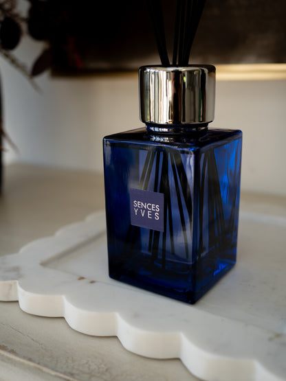 A Sences Yves reed diffuser with a faceted cobalt blue glass bottle, a shiny silver metal collar, and black reeds, resting on a white scalloped tray.