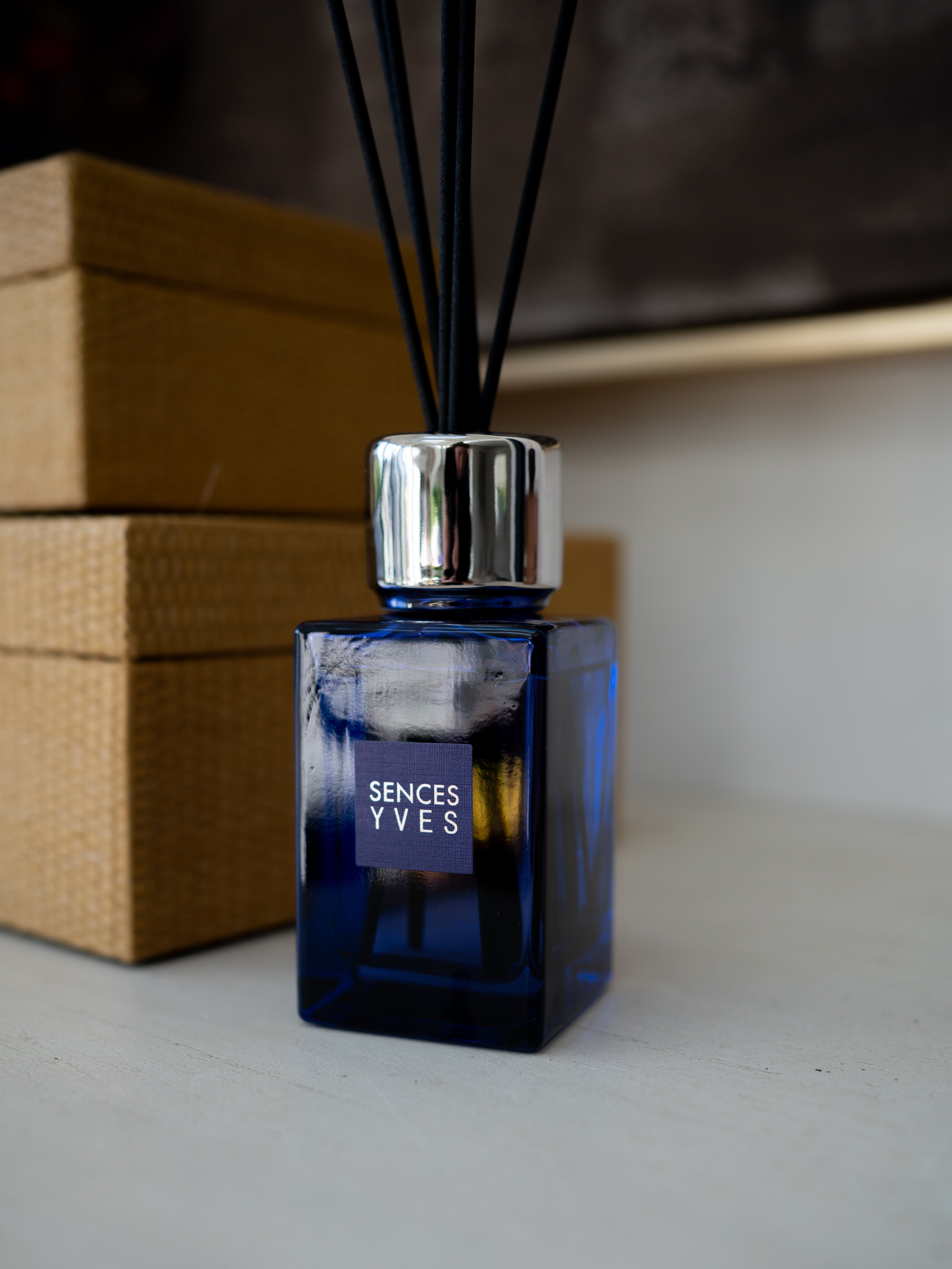 A SENCES YVES reed diffuser with a deep cobalt blue glass bottle, a shiny silver metal collar, and black reeds, set on a light surface with a brown woven box in the background.