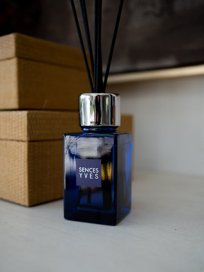 A SENCES YVES reed diffuser with a deep cobalt blue glass bottle, a shiny silver metal collar, and black reeds, set on a light surface with a brown woven box in the background.
