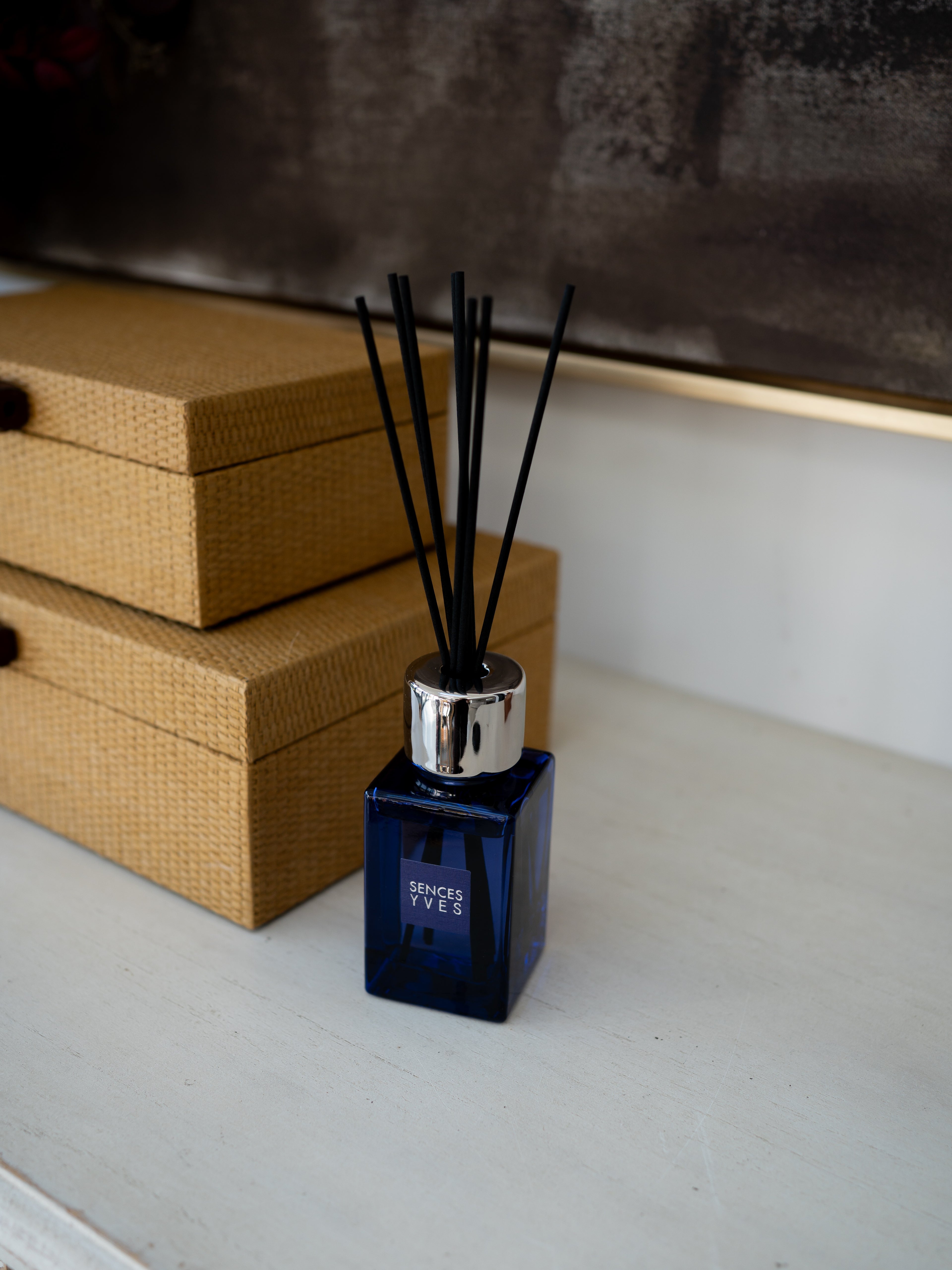 A reed diffuser bottle with a dark blue glass body, silver cap, and black reeds, placed on a light surface next to two tan woven boxes.