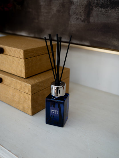 A reed diffuser bottle with a dark blue glass body, silver cap, and black reeds, placed on a light surface next to two tan woven boxes.