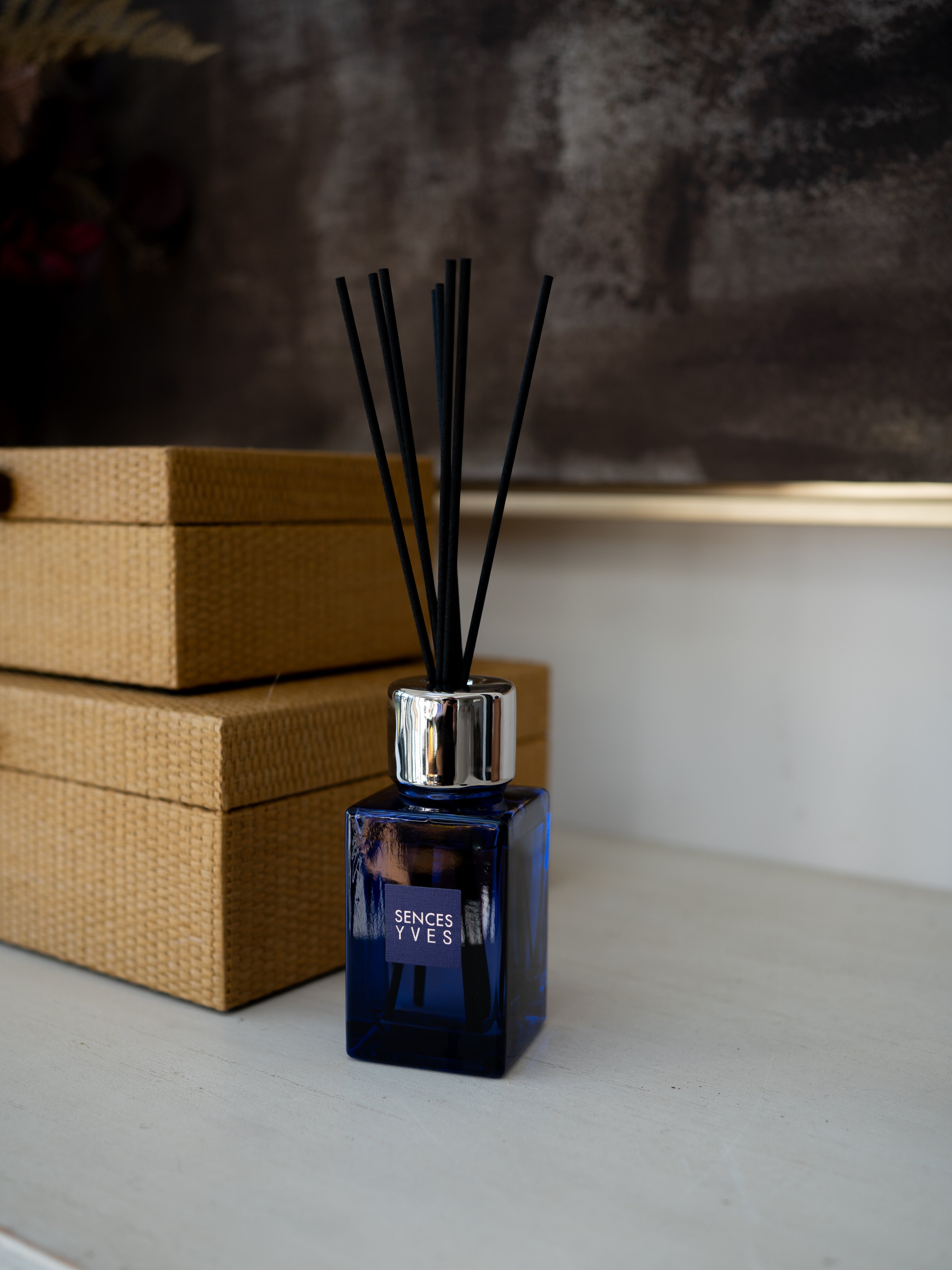 A blue glass reed diffuser bottle with black diffuser sticks and a silver cap, placed on a light surface next to two woven boxes. The bottle is labeled &