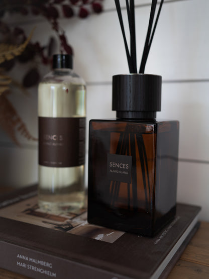 A Sences alang alang room diffuser bottle with black reeds in a dark amber glass bottle, displayed next to a matching Sences diffuser liquid bottle on a book.