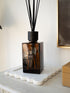 An amber sences glass extra large reed diffuser bottle with black reeds and a black cap, displayed on a white scalloped tray on a light-colored table against a white wall.