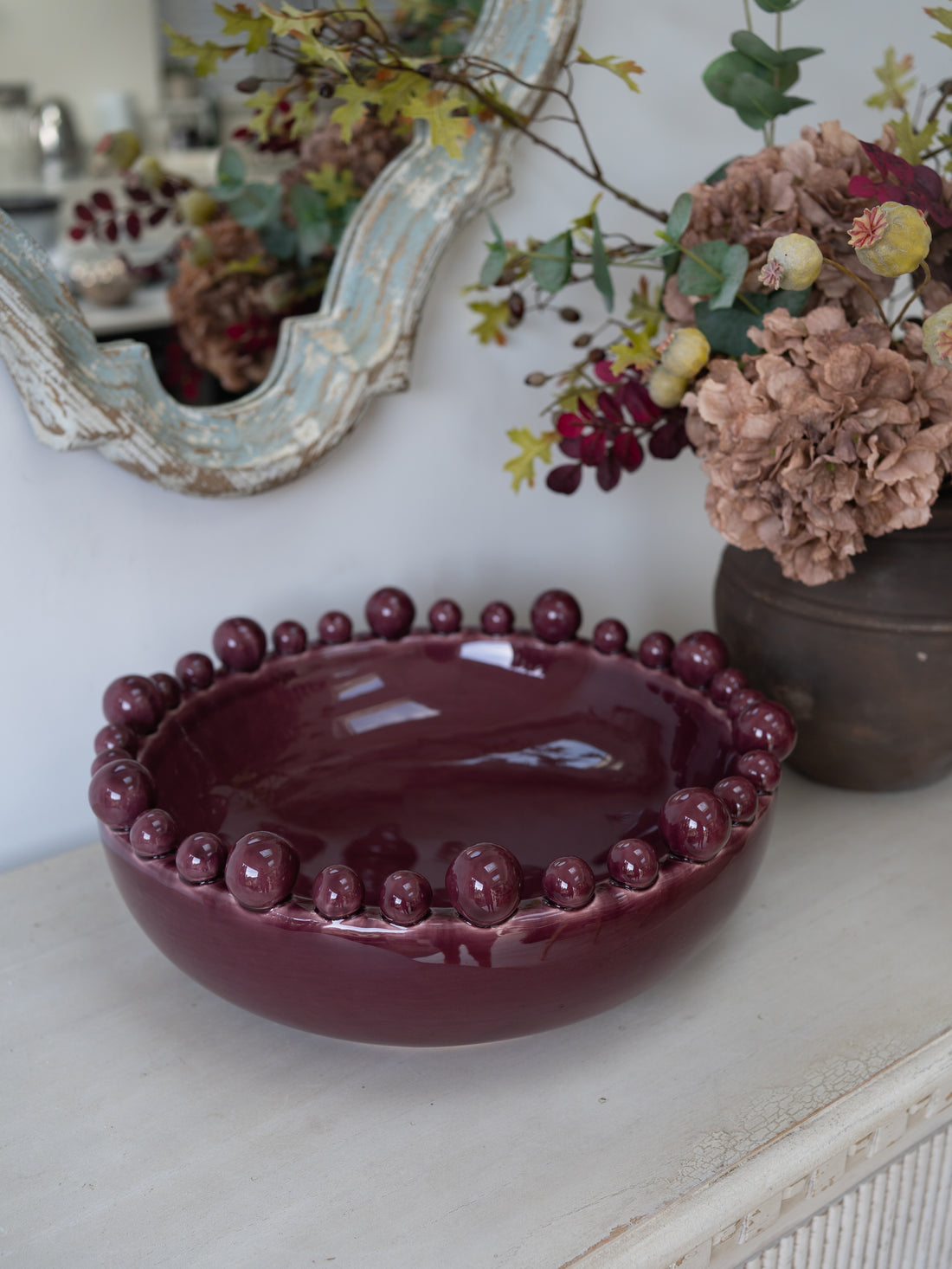 A glossy deep burgundy ceramic bowl with a beaded rim, displayed on a light-colored console table next to a mirror and a vase of faux autumn flowers.