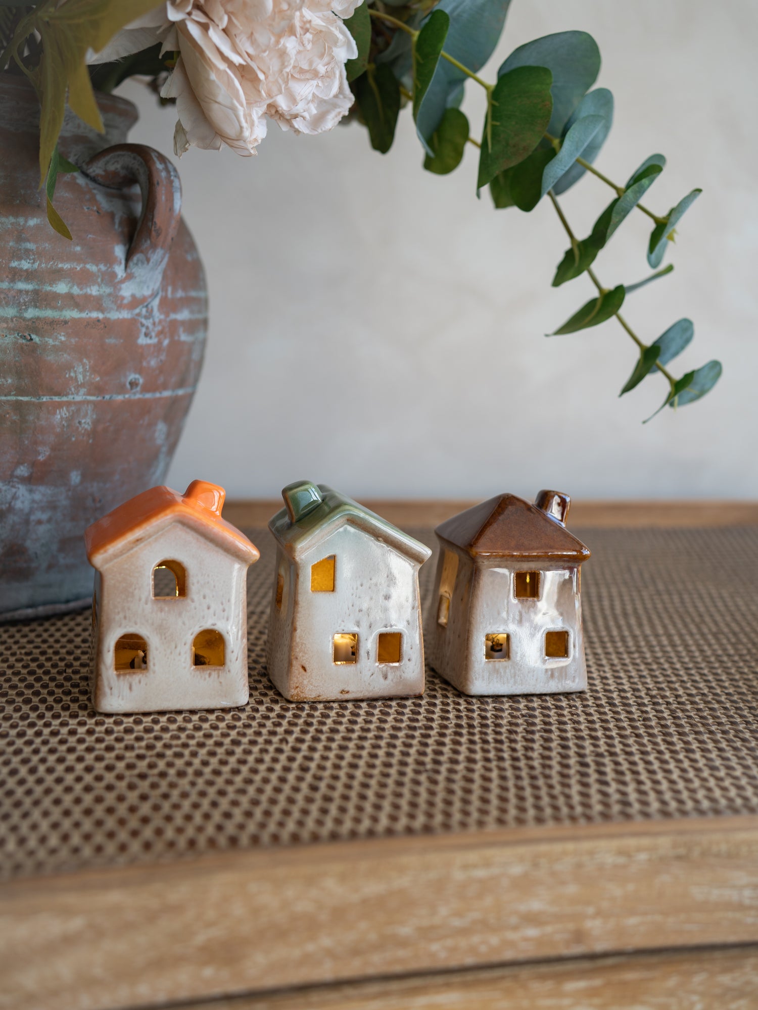 Three small ceramic house-shaped ornaments with orange, green, and brown roofs, displayed on a woven brown mat with a rustic vase and greenery in the background.