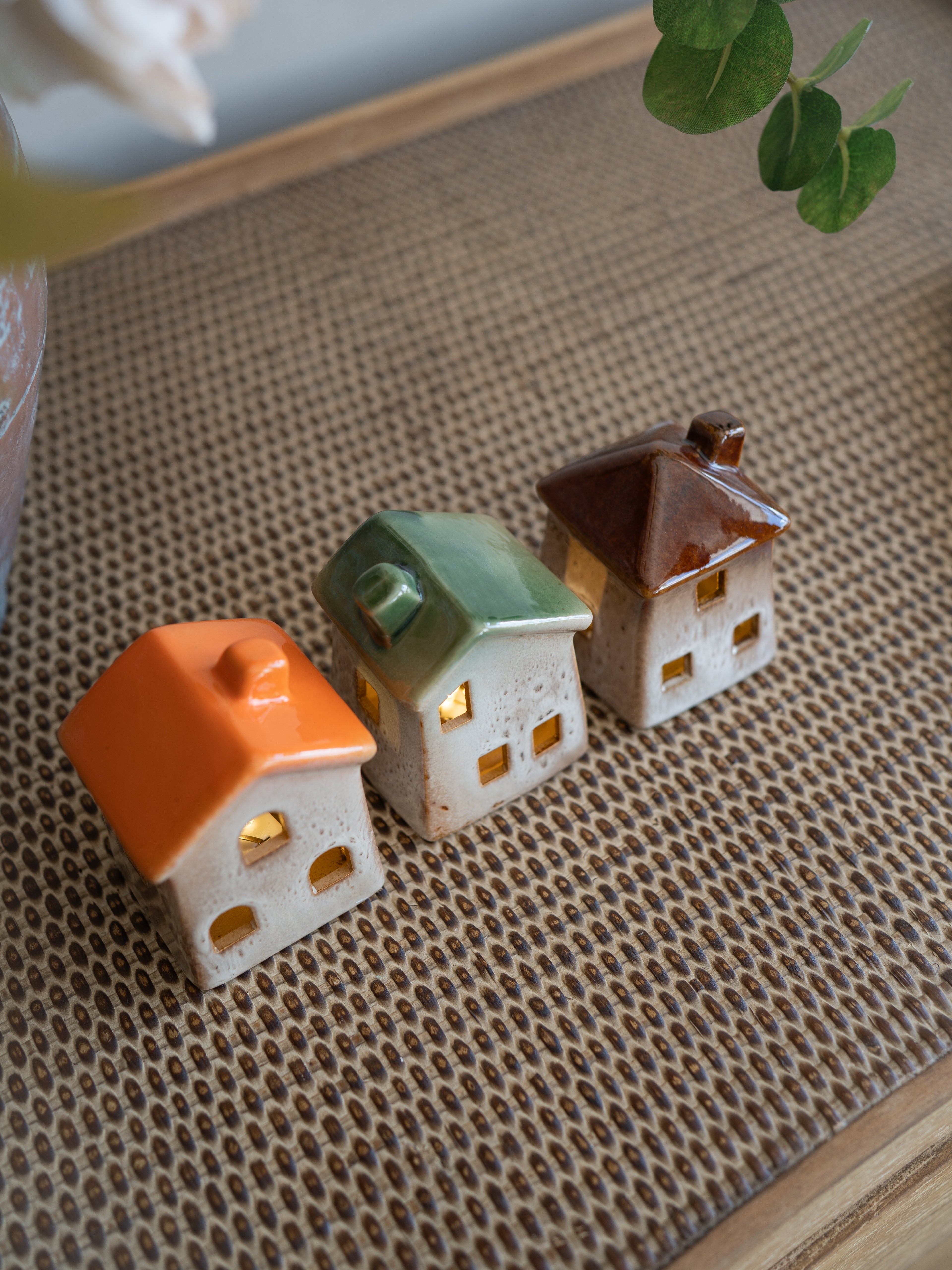 Three small ceramic house-shaped with warm yellow lights inside, arranged on a woven beige mat with a blurred green leaf in the background.