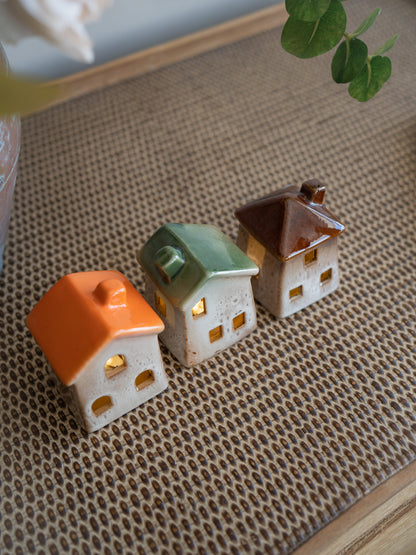 Three small ceramic house-shaped with warm yellow lights inside, arranged on a woven beige mat with a blurred green leaf in the background.