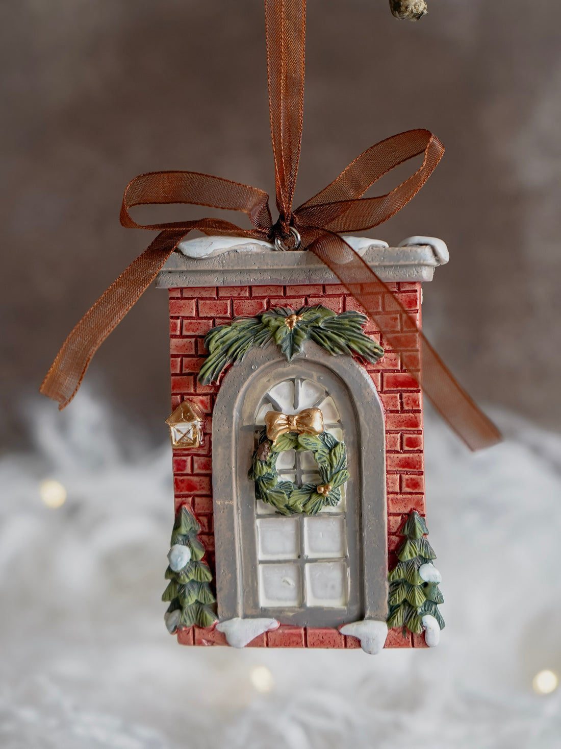 Decorative ornament resembling a brick building with a wreath, hanging from a ribbon against a blurred snowy background.