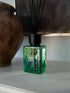 A green glass reed diffuser bottle with black reeds and a black cap, labeled &