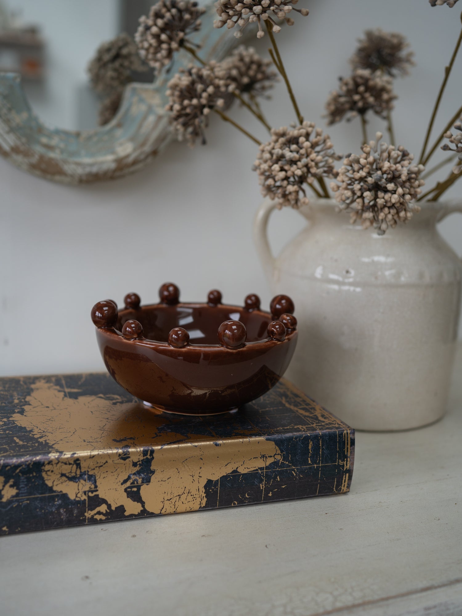 A small glossy brown ceramic bowl with raised spherical details around the rim, displayed on a dark blue and gold book in front of a white ceramic vase with dried flowers.